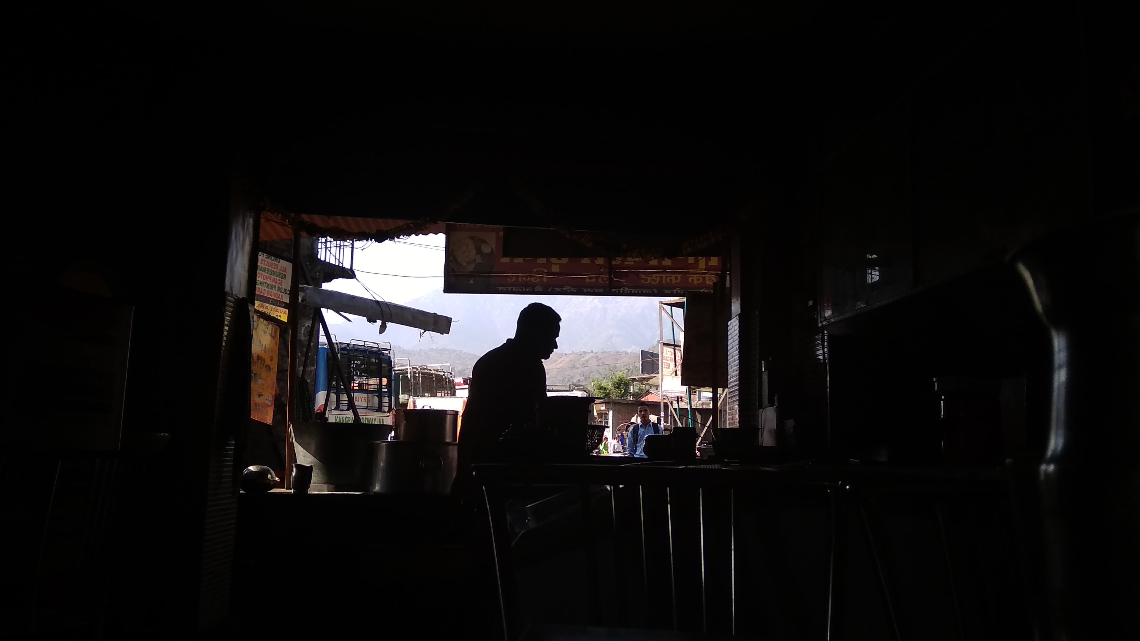 A cook silhouetted in the open doorway of a dhaba in Palampur — Hindi signage overhead, mountains visible beyond.